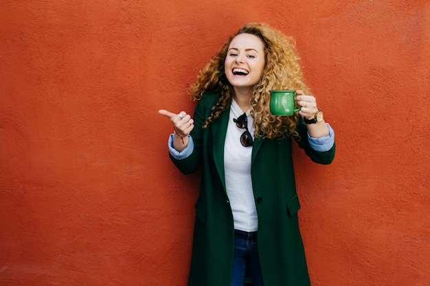 Excited woman with curly blonde hair wearing jacket holding green cup of coffee raising her thumb.