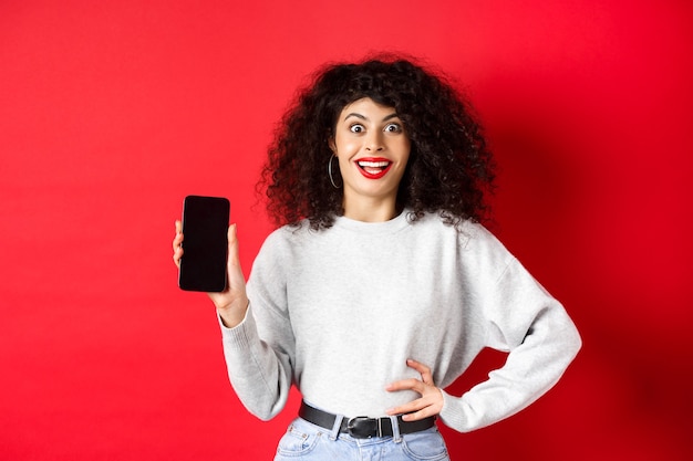 Excited woman with curly hair and red lips, showing empty smartphone screen and screaming from joy, standing on red wall.