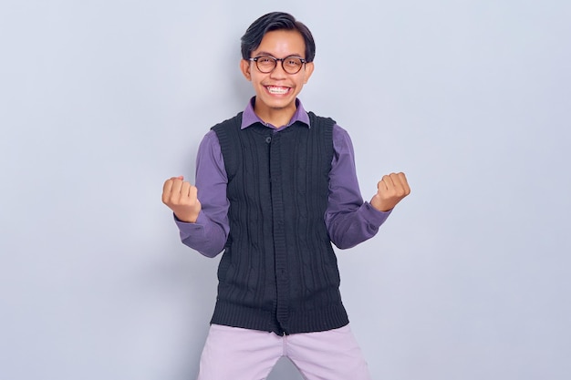 Excited young Asian man in casual shirt and vest celebrating victory or success with raised fists up isolated on white background People lifestyle concept