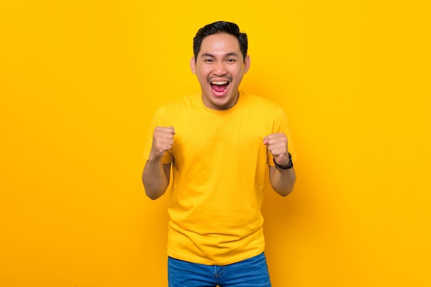 Excited young Asian man in casual tshirt celebrating success with raised fist isolated on yellow background People lifestyle concept