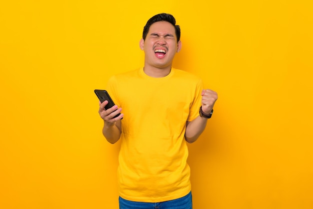 Excited young Asian man in casual tshirt holding mobile phone and making winner gesture getting good news isolated on yellow background People lifestyle concept