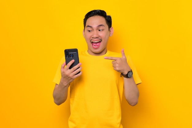 Excited young Asian man in casual tshirt pointing fingers at mobile phone reacting to new app isolated on yellow background People lifestyle concept