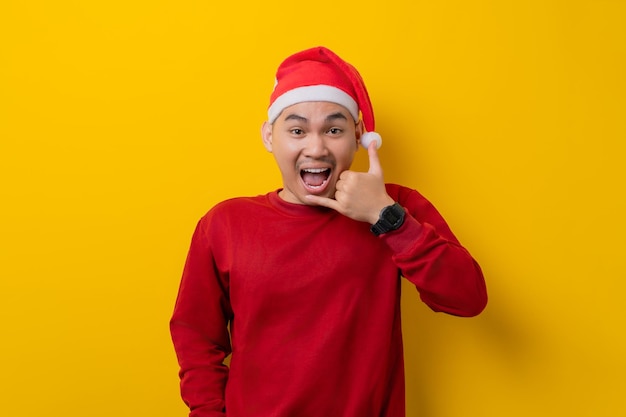 Excited young Asian man in Santa hat doing phone gesture call me back on yellow studio background celebration Christmas holiday and New Year concept