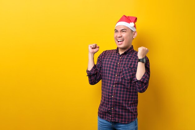 Excited young Asian man in Santa hat raising hands up celebrating success on yellow studio background Happy New Year 2023 celebration merry holiday concept