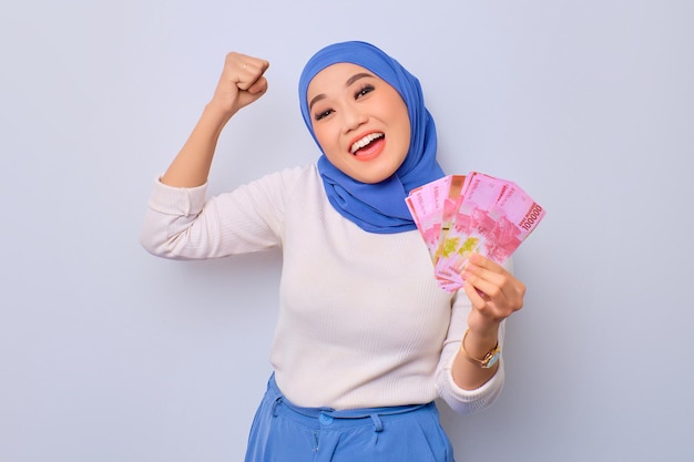 Excited young Asian Muslim woman holding money banknotes and celebrating success isolated over white background