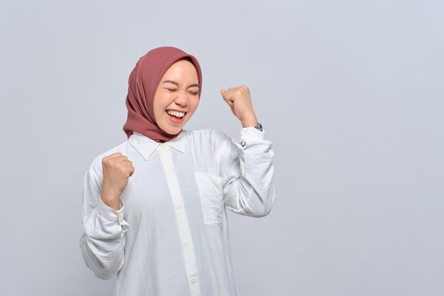 Excited young Asian Muslim woman raising fist up celebrating success isolated over white background