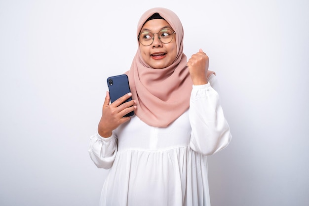 Excited young Asian Muslim woman using a mobile phone and celebrating success getting good news