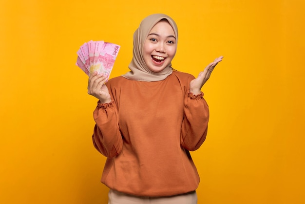 Excited young Asian woman in orange shirt holding money banknotes isolated over yellow background