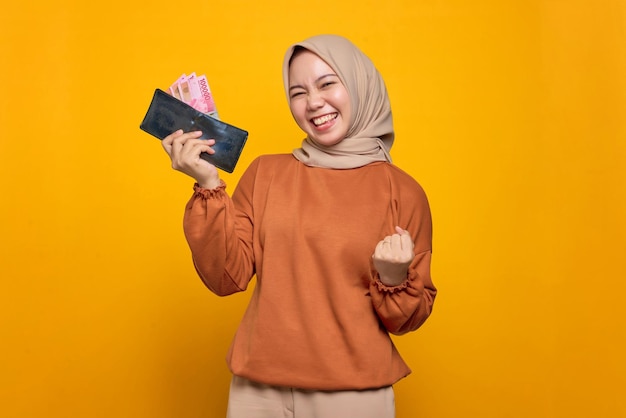 Excited young Asian woman in orange shirt holding wallet full of money banknotes isolated over yellow background