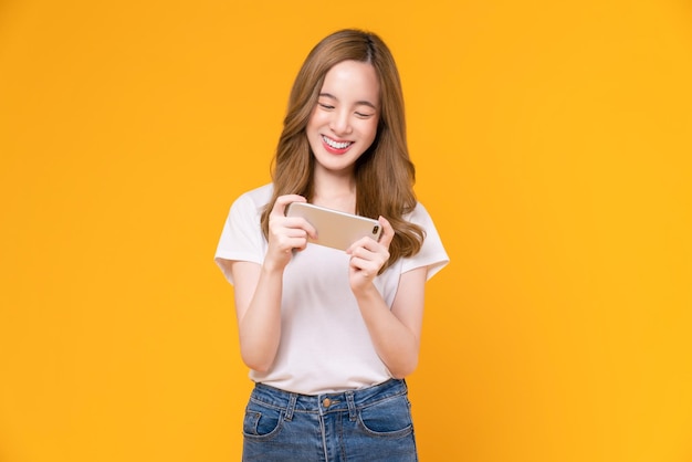 Excited young Asian woman playing games with smartphone on orange background.