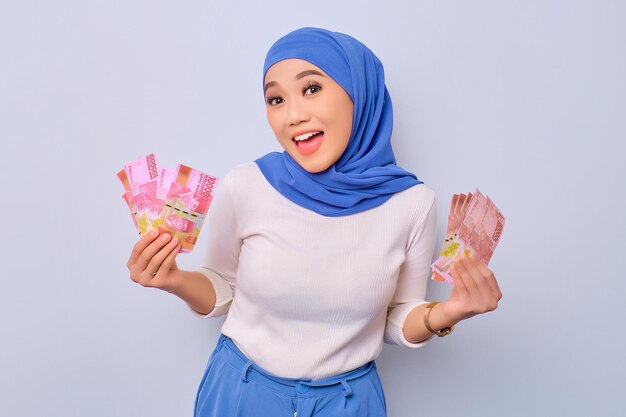 Excited young beautiful Asian Muslim woman holding money banknotes isolated over white background