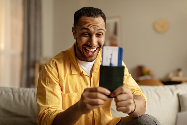 Excited young black man with passport and tickets getting ready for summer vacation sitting on couch