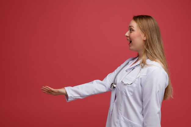 Excited young blonde female doctor wearing medical robe and stethoscope around neck standing in profile view looking at side stretching out hand 