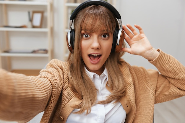 Excited young blonde student lady freelancer in warm sweater in headphones listen fav songs sitting in armchair at modern home interior Music time Relaxing Cool playlist Concept Wide selfie view