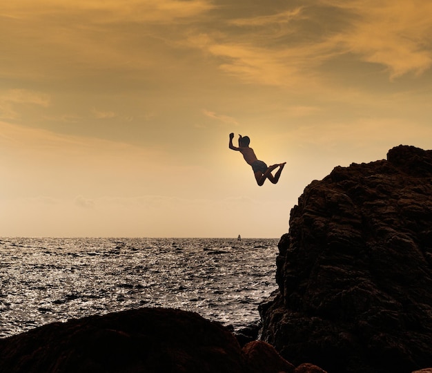 Excited young boy jumping from a rocky cliff into the lake water surrounded by beautiful sunset view