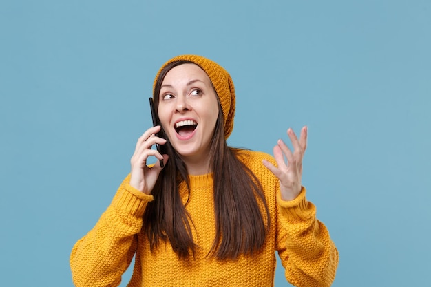 Excited young brunette woman girl in yellow sweater and hat posing isolated on blue background studio portrait. People sincere emotions lifestyle concept. Mock up copy space. Talking on mobile phone.