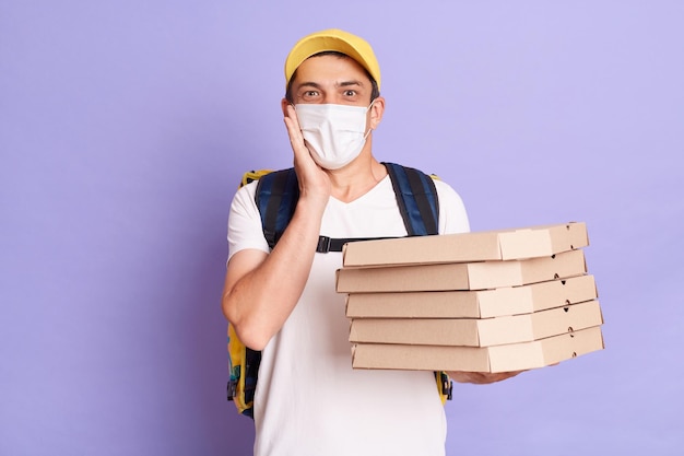 Excited young Caucasian delivery man in protective mask holding pizza boxes isolated on purple background looking at camera with amazement keeps hand on cheek looking at camera