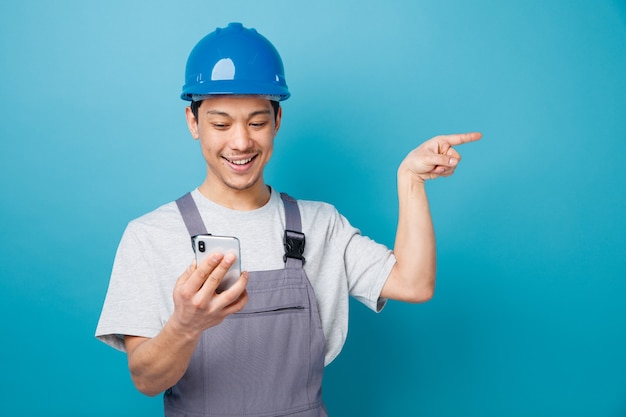 Excited young construction worker wearing safety helmet and uniform holding and looking at mobile phone pointing to side 