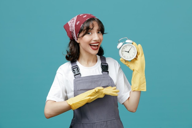 Excited young female cleaner wearing uniform bandana and rubber gloves showing alarm clock pointing at it with hand looking at camera isolated on blue background