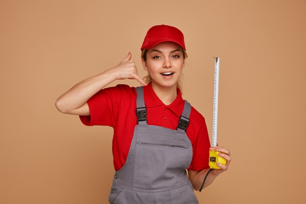 Excited young female construction worker wearing cap and uniform holding tape meter doing call gesture 