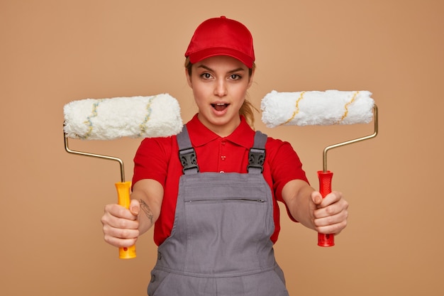 Excited young female construction worker wearing cap and uniform stretching out paint rollers towards camera 