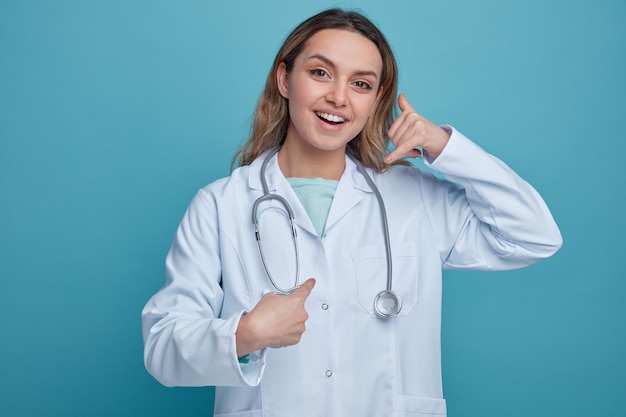 Excited young female doctor wearing medical robe and stethoscope around neck doing call gesture pointing at herself 