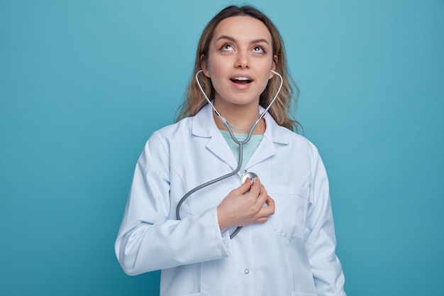 Excited young female doctor wearing medical robe and stethoscope looking up listening to her own heartbeats 