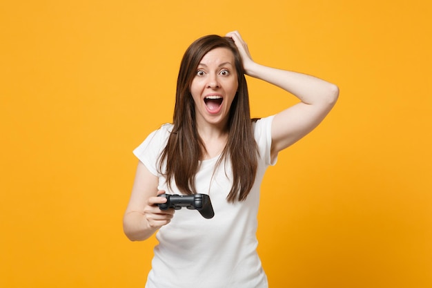 Excited young girl in white casual clothes keeping mouth open, putting hand on head playing video game with joystick isolated on yellow orange background. People lifestyle concept. Mock up copy space.