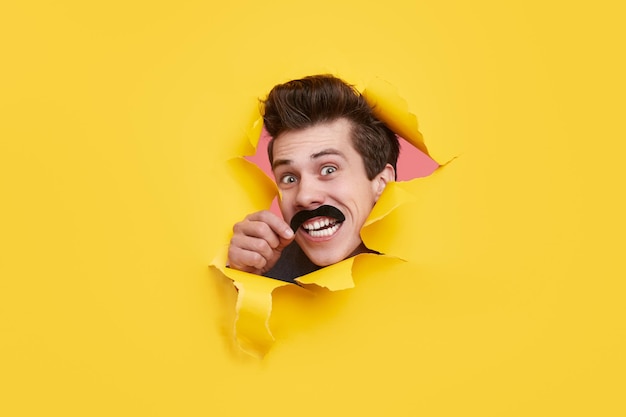 Excited young guy with fake mustache looking at camera with astonished face expression through ripped yellow paper