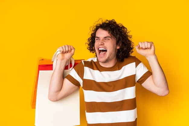 Excited young guy with shopping bags shouting happily in yellow studio