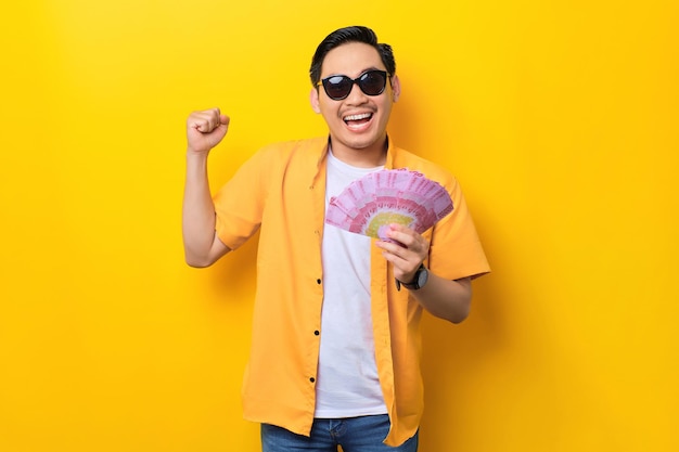 Excited young handsome Asian man holding money banknotes and celebrating success isolated on yellow background