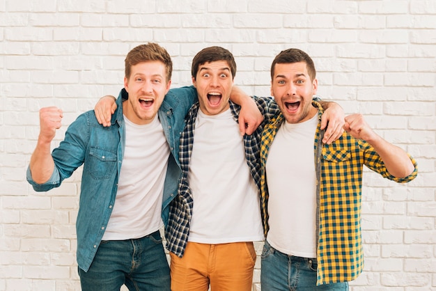 Excited young male friends standing against white wall clenching their fist