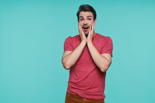 Excited young man in casual clothing looking at camera and holding hands on face