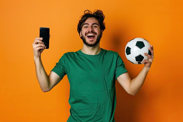 Excited young man in green shirt supports his favorite football team with soccer ball and blank phon