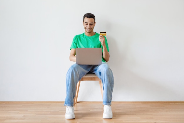 Excited young man holding credit card and laptop sitting on chair buying things or ordering goods in web store