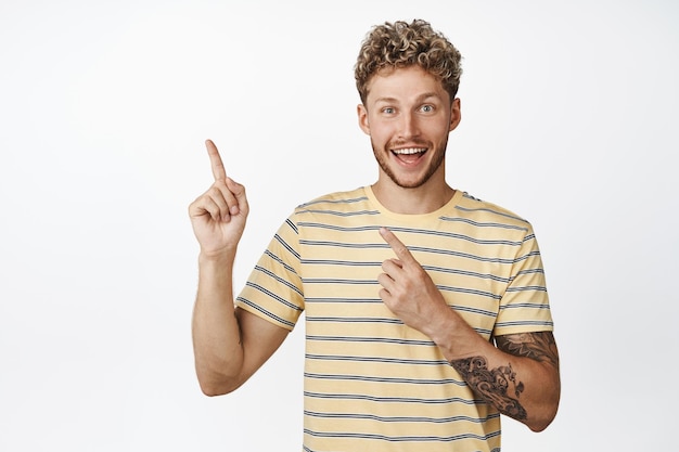 Excited young man showing announcement smiling from big amazing news pointing at upper left corner standing over white background