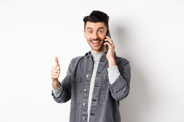 Excited young man talking on phone and showing thumbs up, smiling satisfied, standing against white background.