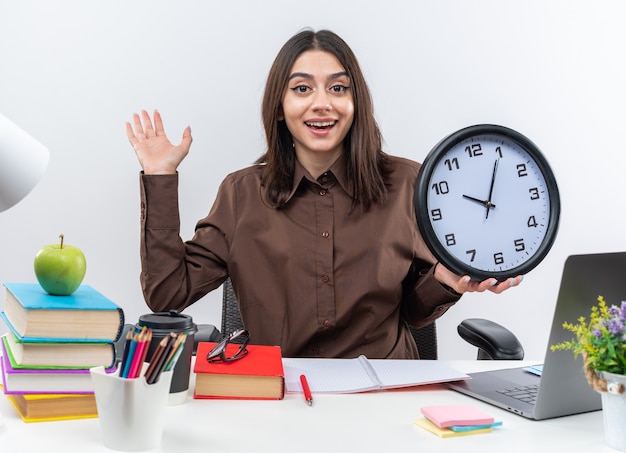 Excited young school woman sits at table with school tools holding wall clock 