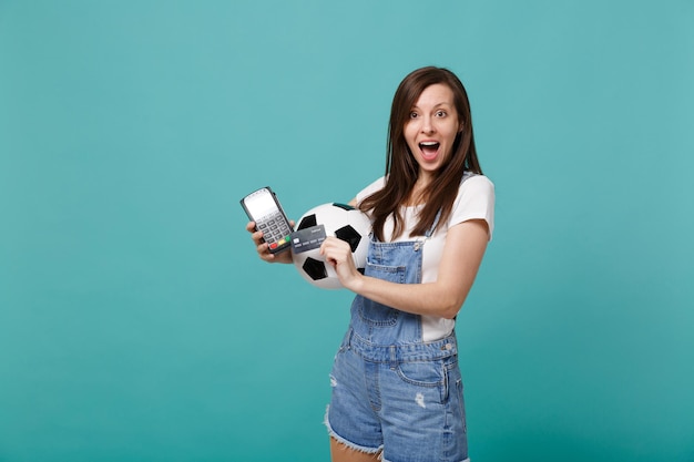 Excited young woman football fan support favorite team with soccer ball, wireless modern bank payment terminal to process and acquire credit card payments isolated on blue turquoise wall background.