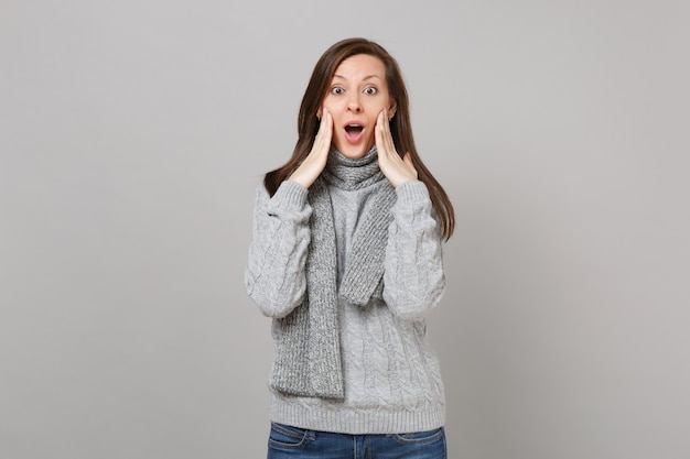 Excited young woman in gray sweater, scarf keeping mouth wide open put hands on cheeks isolated on grey background. Healthy fashion lifestyle, people emotions, cold season concept. Mock up copy space.