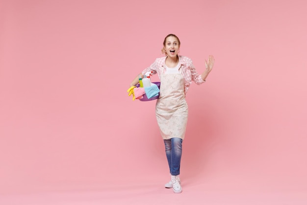 Excited young woman housewife in apron hold basin with detergent bottles washing cleansers while doing housework isolated on pink background studio portrait. Housekeeping concept. Spreading hands.