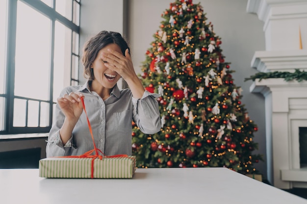 Excited young woman unpacking christmas gift while sitting in room decorated with bright xmas tree