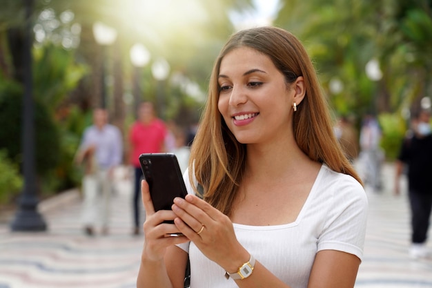 Excited young woman watching her smartphone when walking in the street with blurred people on the background Beautiful girl using mobile app outdoors Teenager lifestyle technology concept