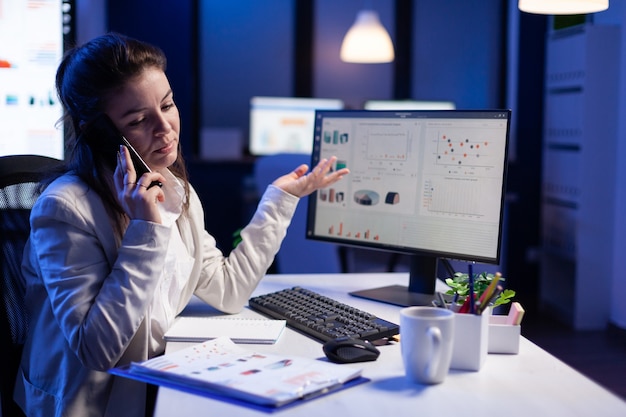 Executive entrepreneur talking at smartphone with employee creates new marketing concept in business office. Busy manager using modern technology network sitting at desk late at night