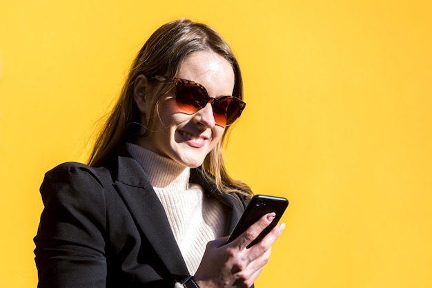 Executive entrepreneur woman wearing black jacket and wool sweater and sunglasses on yellow background in the street with copy space looking at mobile Entrepreneur woman concept