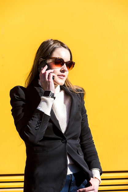 Executive entrepreneur woman wearing black jacket and wool sweater and sunglasses on yellow background in the street with copy space talking on the mobile Entrepreneur woman concept