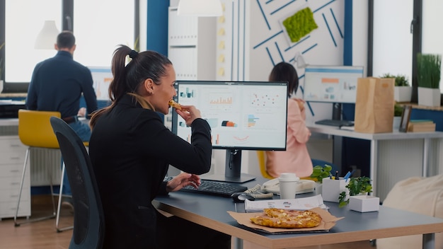 Executive manager eating tasty pizza while typing financial statistics at desk in workplace