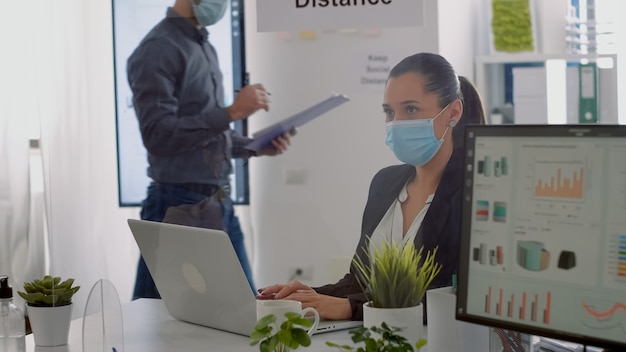 Executive manager with face mask typing marketing information on laptop computer while sitting at desk table in company office. Team respecting social distance to avoid infection with covid19