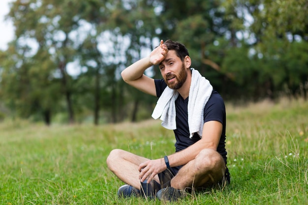 Exhausted after workout training or jogging athlete man sitting outdoors with a towel. Portrait sweaty fitness male in city park background forest and trees. Caucasian sportsman resting after running