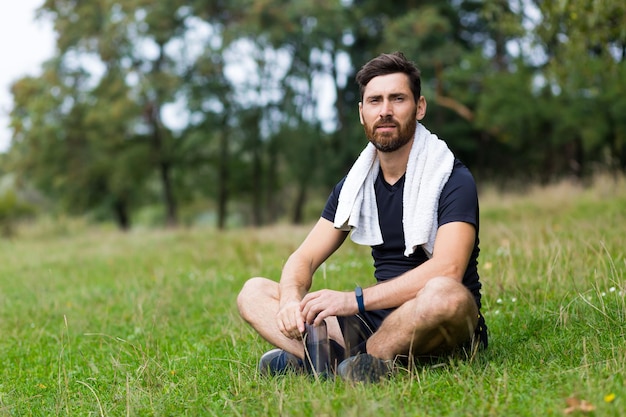 Exhausted after workout training or jogging athlete man sitting outdoors with a towel. Portrait sweaty fitness male in city park background forest and trees. Caucasian sportsman resting after running